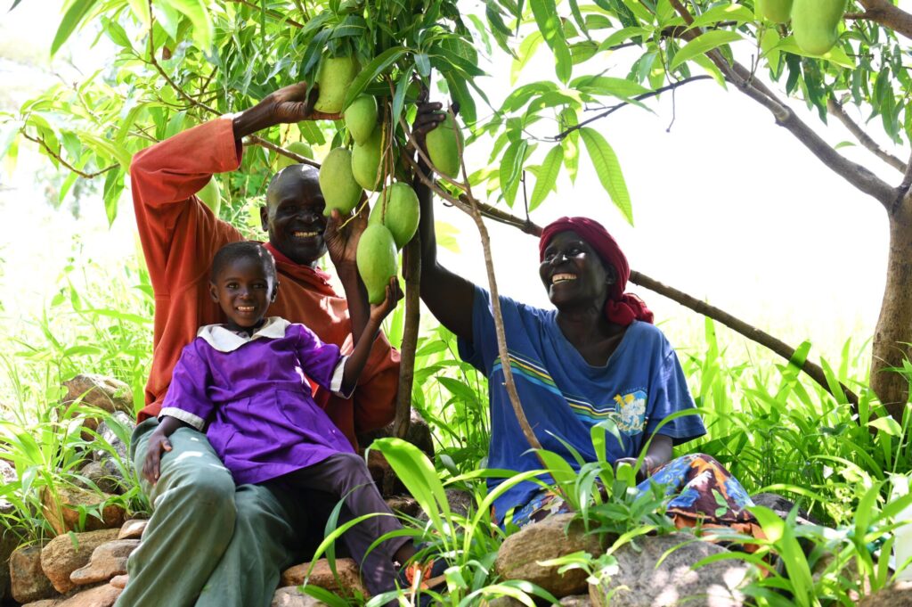 Joyce, Edward ja Blessing istumassa ja hymyilemässä mangopuun alla.