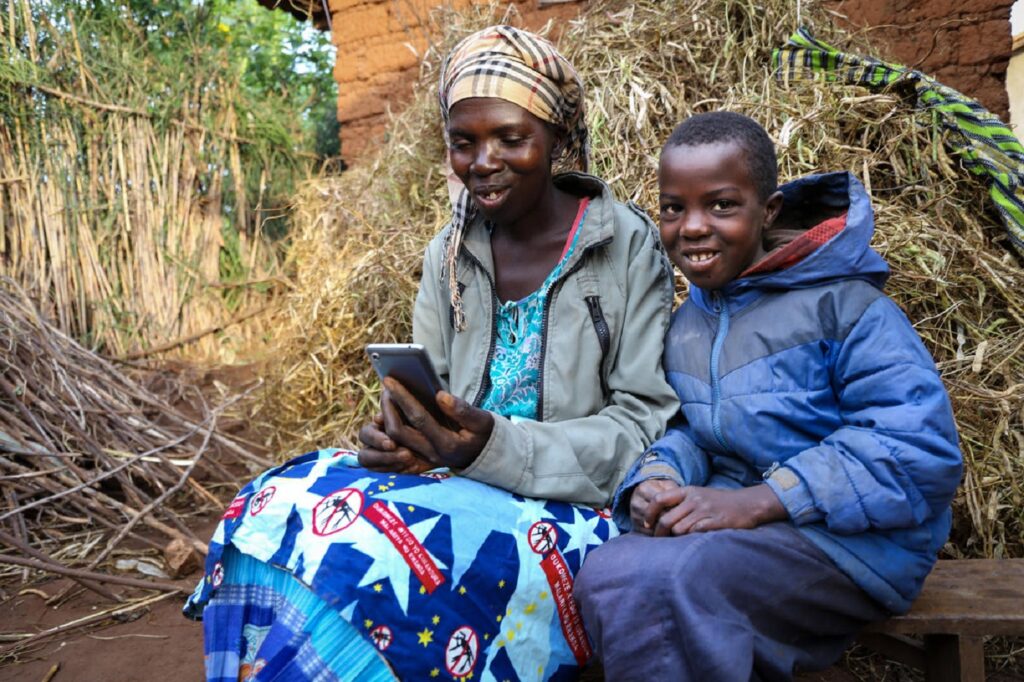 Ruandalainen äiti ja poika istuvat ulkona penkillä. Äiti katsoo kädessä pitävänsä kännykkää ja poika katsoo kameraan. Molemmat hymyilevät.