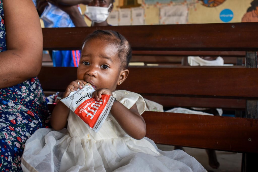 Valkoiseen mekkoon pukeutunut Etelä-Sudanilainen pikkutyttö syö maapähkinätahnaa hätäruokapussista ja katsoo kameraan.