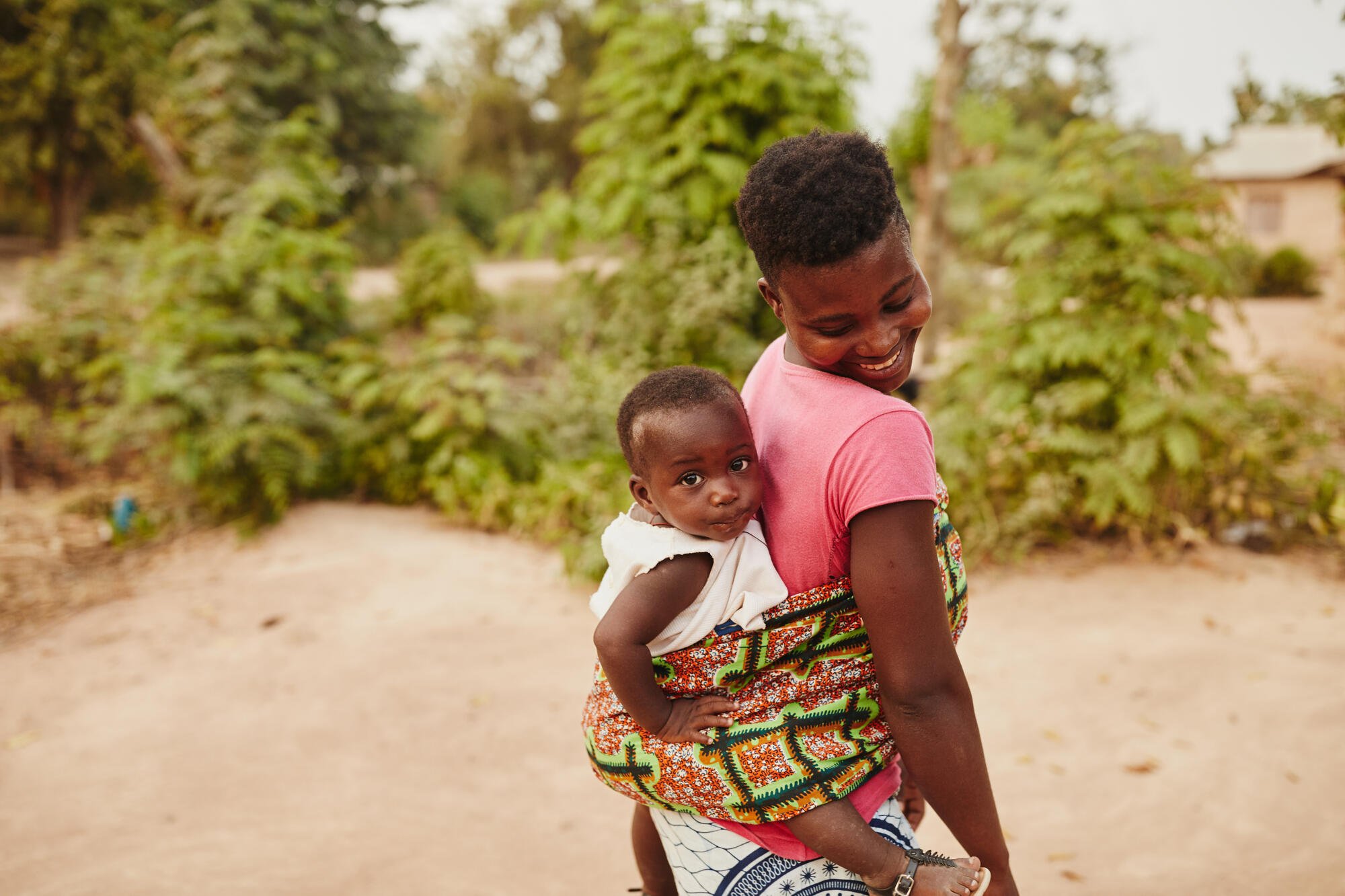 Ghanalainen äiti katsoo hymyillen taaksepäin, selällään kantavansa taaperoaan, joka katsoo itsevarmasti kameraan.