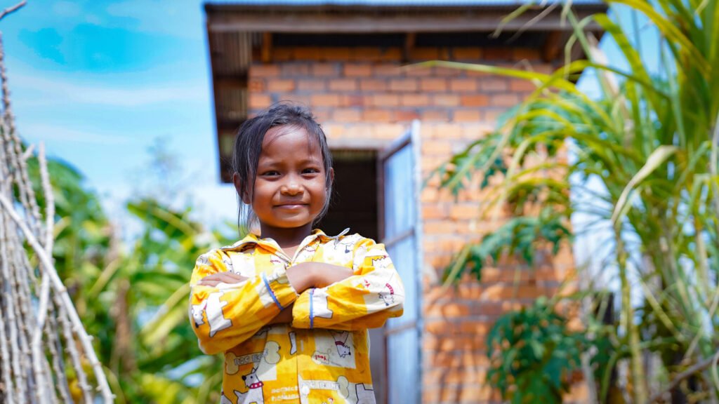 Kambodzalainen pikkutyttö seisoo kädet ristissä pienen rakennuksen ulkopuolella ja hymyilee itsevarmasti kameralle.