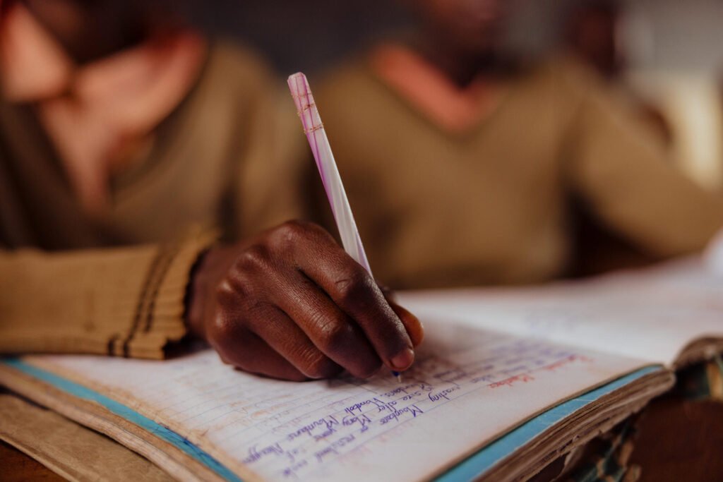Lähivkuva kenialaisen tytön kädestä, joka pitää kynää ja kirjoittaa kouluvihkoon.