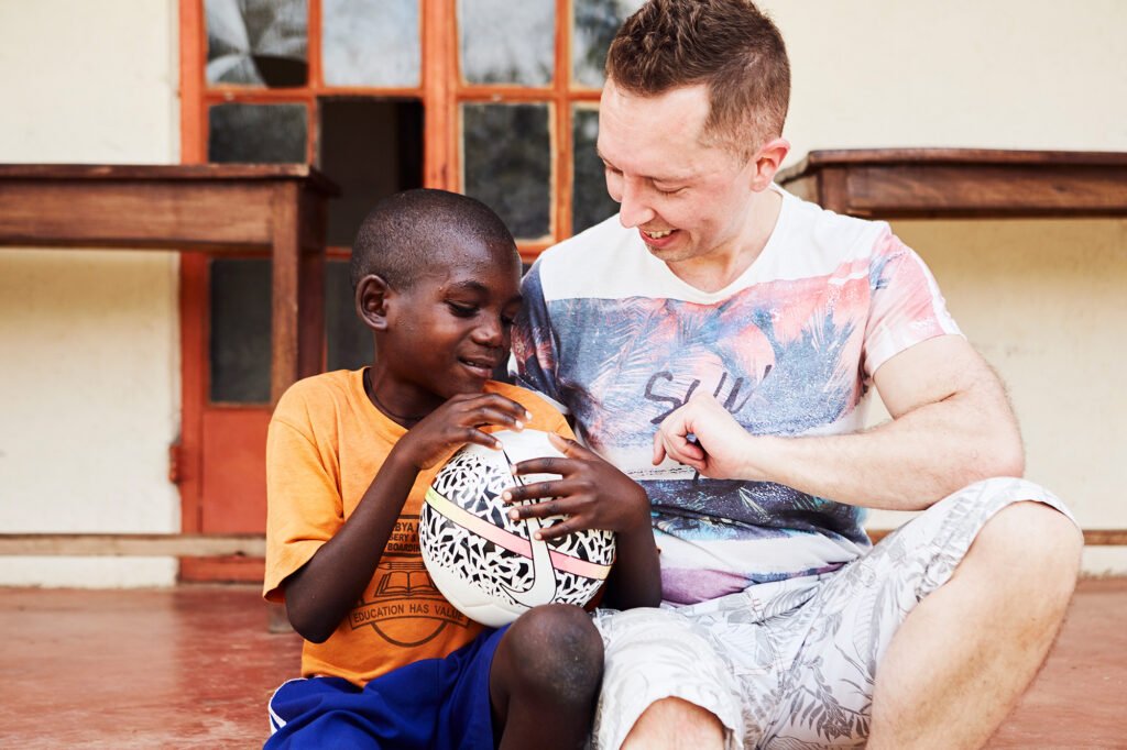 Suomalainen kummi. Mika, istuu ugandalaisen kummilapsensa vieressä Ugandassa. Kummilapsella on sylissä jalkapallo ja molemmat katsovat sitä hymyillen.