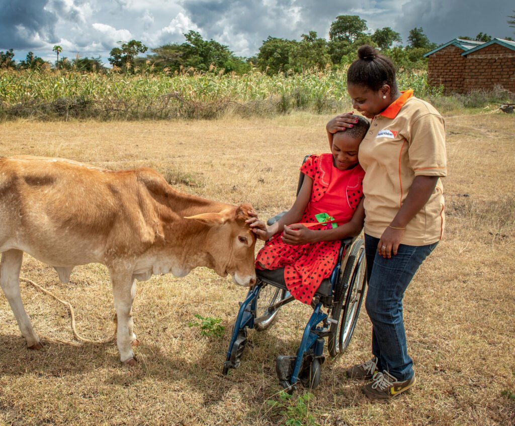 Pyörätuolissa istuva tyttö silittää lehmän päätä ja nojaa World Visionin paikallista työntekijää vastaan.