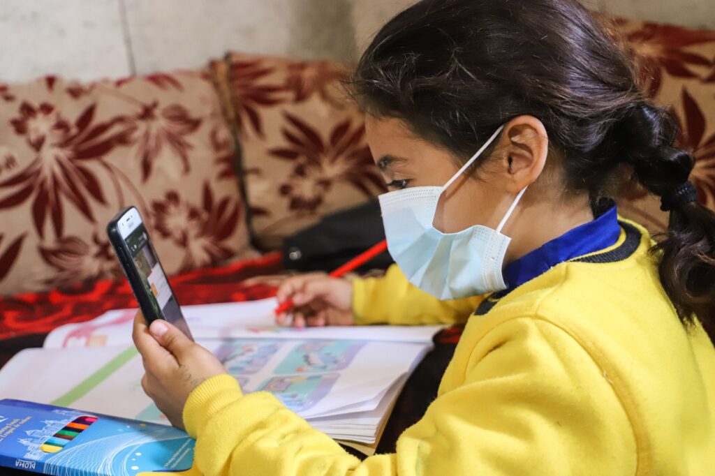 Keltaiseen pukeutunut libanonilainen tyttö katsoo kännykältä koulutehtäviään ja kirjoittaa samalla vihkoon.