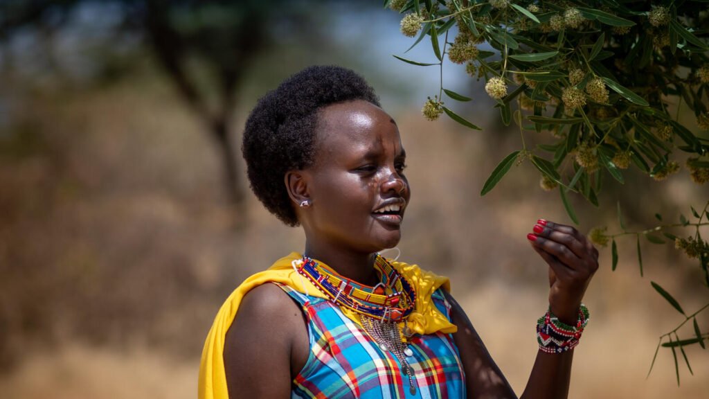 Värikkäisiin vaatteisiin pukeutunut Nancy Yiampoi seisoo puun varjossa Keniassa.