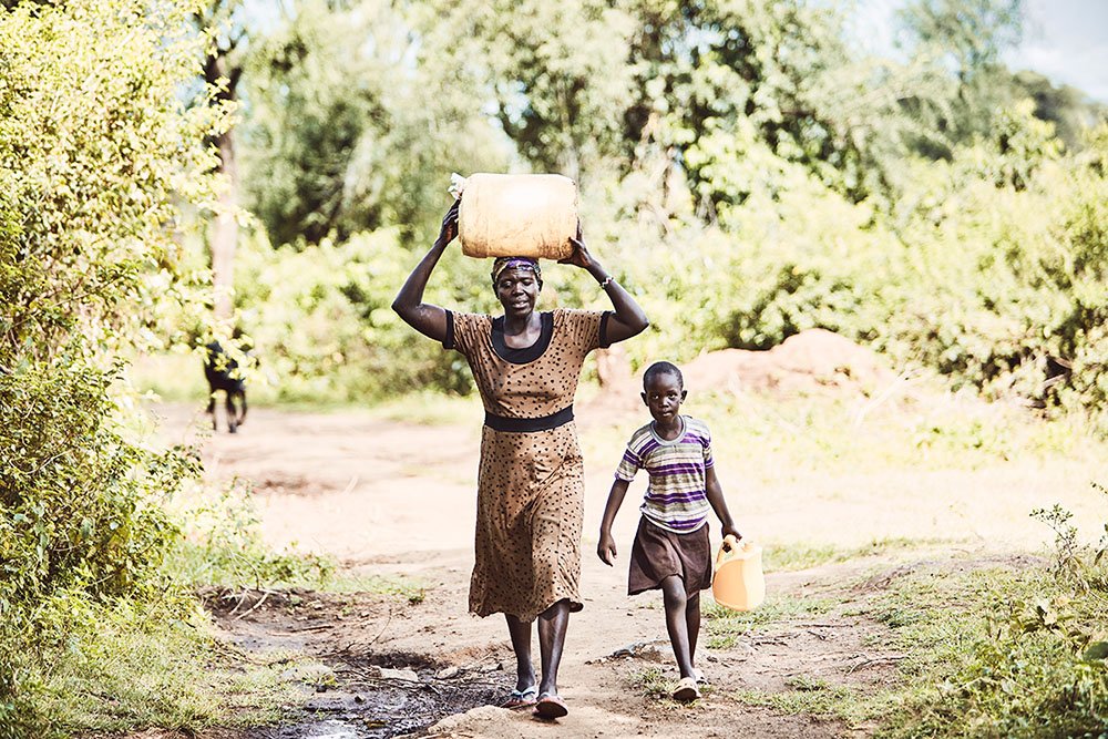 Ngoswetilainen äiti kantamassa tavaraa pään päällä. Vieressä kävelee lapsi kantamassa vesikanisteria.