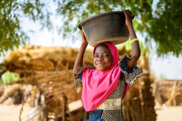 Nigerilainen nuori tyttö kantaa vettä astiassa pään päällä ja hymyilee itsevarmasti kameralle.