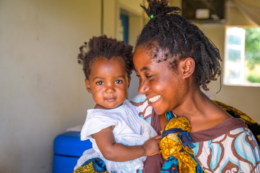 Sambialainen äiti pitää taaperoaan ja hymyilee leveästi hänelle. Taapero katsoo iloisesti kameraan.