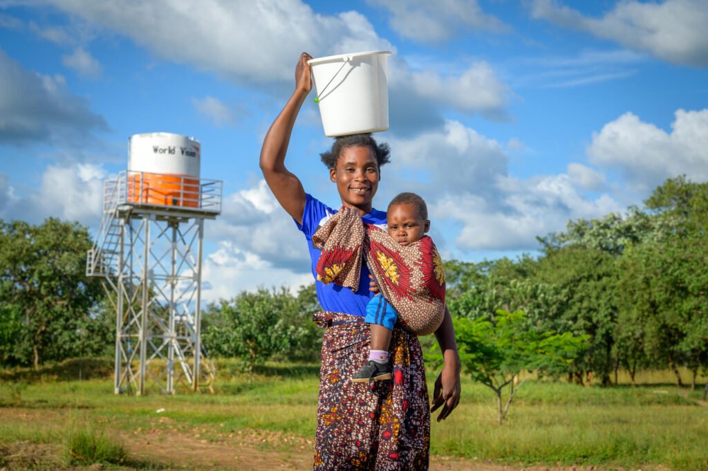 Sambialainen Violet poseeraa hymyillen kameralle. Päällään hän kantaa ämpärillisen puhdasta vettä. Hän kantaa taaperonsa, Daliso, kantoliinassa.