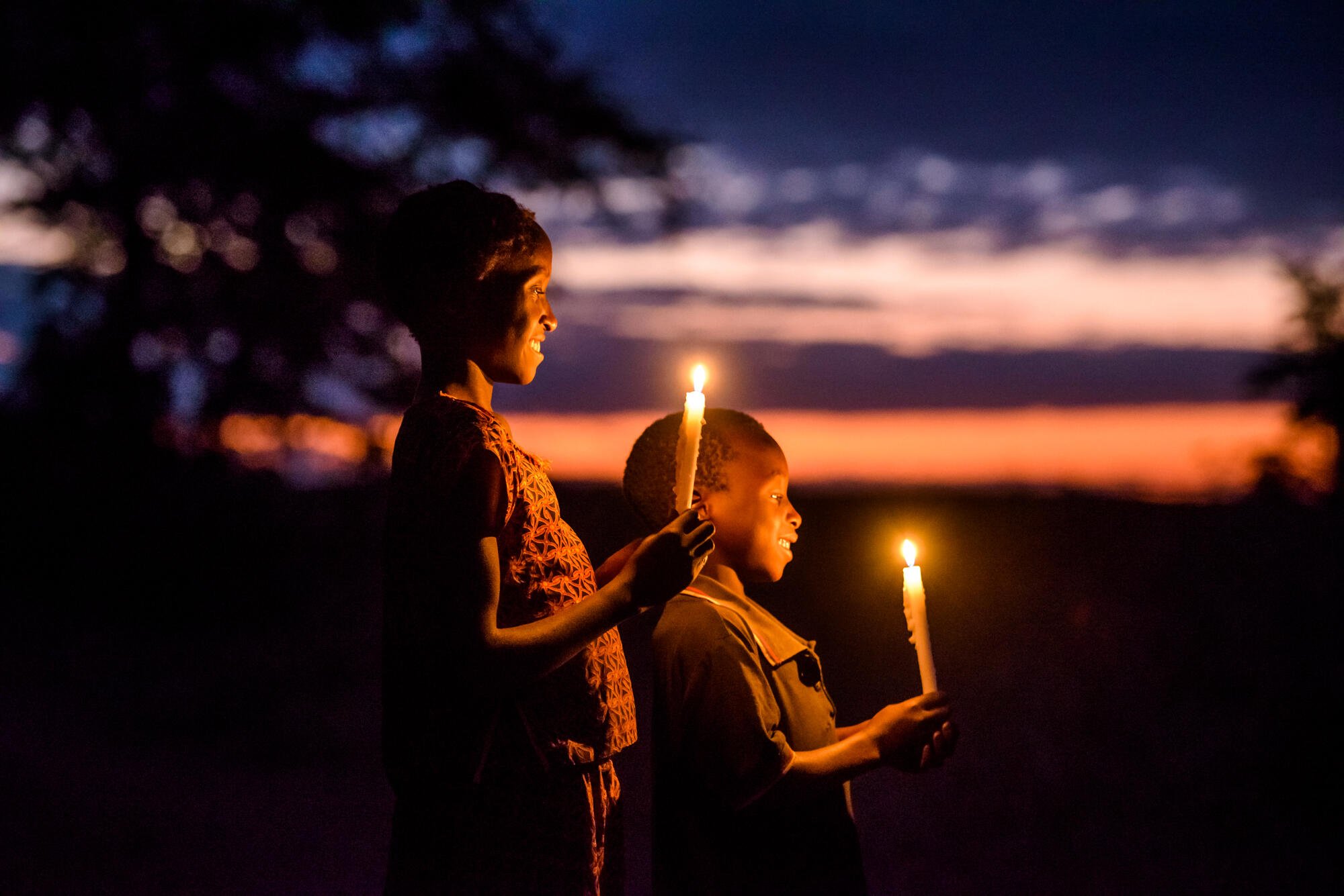 Kaksi Sambialaista lasta pitävät kynttilöitä ja hymyilevät. Takana näkyy auringonlasku.