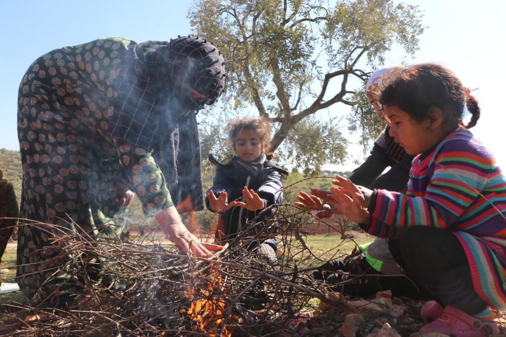 Syyrialaisia lapsia lämmittämässä kädet nuotion ääressä. Aikuinen nainen laittaa samalla lisää oksia nuotioon.