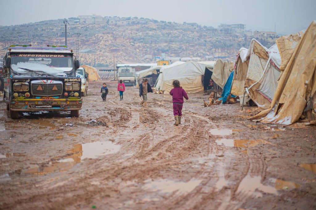 Näkymät Luoteis-Syyrian pakolaisleirillä. Eilaisia telttoja on pystytty hyvin mutaisen tien varteen, joka on täynnä vesilätäköitä. Tiellä kävelee pieniä lapsia.