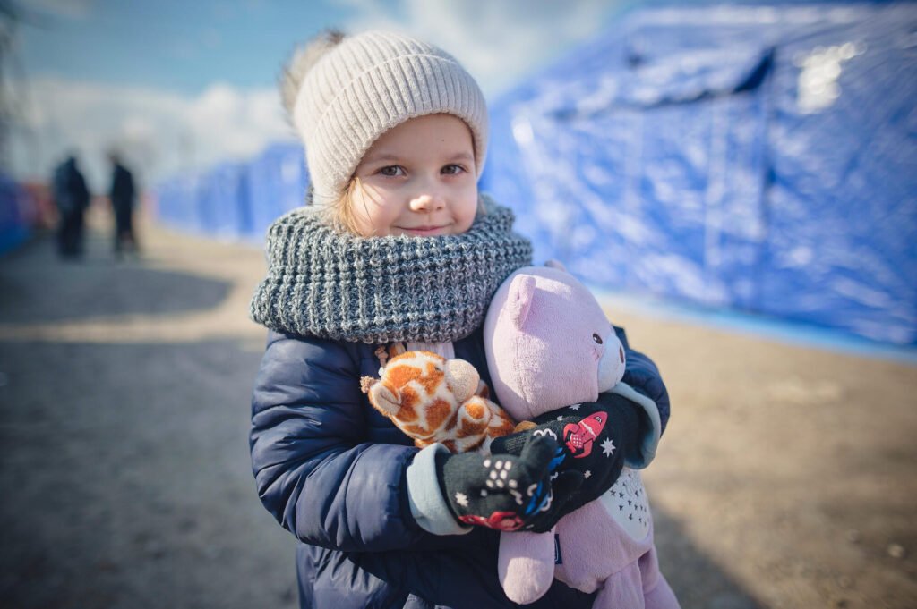 Ukrainalainen pikkutyttö hymyilee ujosti kameralle ja halaa kahta pehmolelua. Taustalla näkyy telttoja.