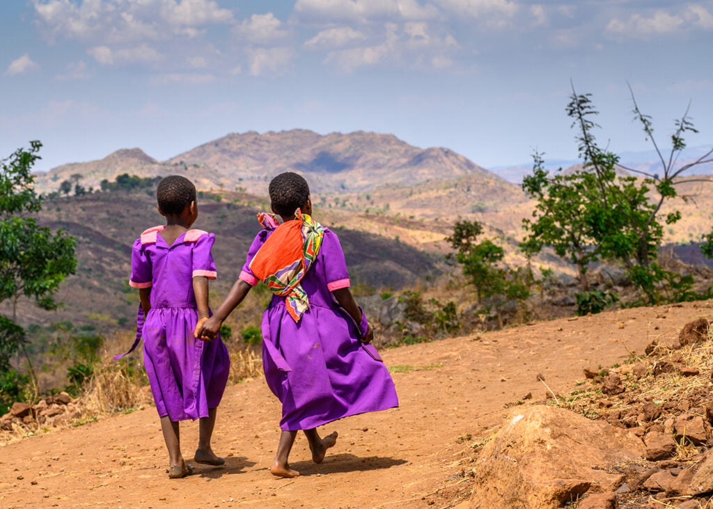 Lapset kävelevät kuivalla aavikolla liilat mekot päällä. Heidät on kuvattu takaapäin.