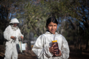 Bolivialainen tyttö pitää hynajapurkkia käsisään ja hymyilee kameralle. Hän on pukeutunut valkoiseen mehiläispitäjän asuun. Taustalla näkyy aikuinen, joka on pukeutunut samanlaiseen asuun.