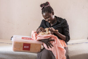 Kenialainen äiti istuu sairaalavuoteella ja hymyilee sylissä olevalle vastasyntyneelle vauvalleen. Hänen vieressä, sängyn päällä, näkyy World Visionilta saatu äitiyspakkaus.