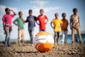 Kuvan edustalla on World Visionin logolla varustettu oranssi-valkoinen jalkapallo. Taustalla näkyy lapsia.