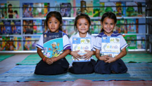 Kolme kambodzalaista koulutyttöä poseeraavat hymyillen kameralle, kirjoja käsissään.