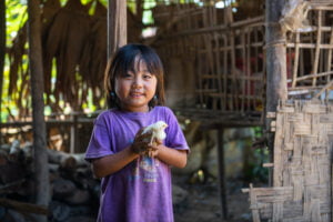 Kambodzalainen pikkutyttö pitää tipua käsissään ja hymyilee kameralle. Taustalla näkyy perheen talo.