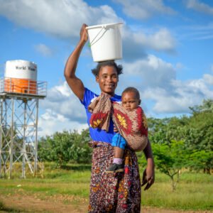 Äiti kantaa vettä sangossa pään päällä ja hymyilee kameralle. Hän pitää taaperonsa kantoliinassa ja taustalla näkyy iso vesitankki, jonka kyljessä on World Visionin logo.