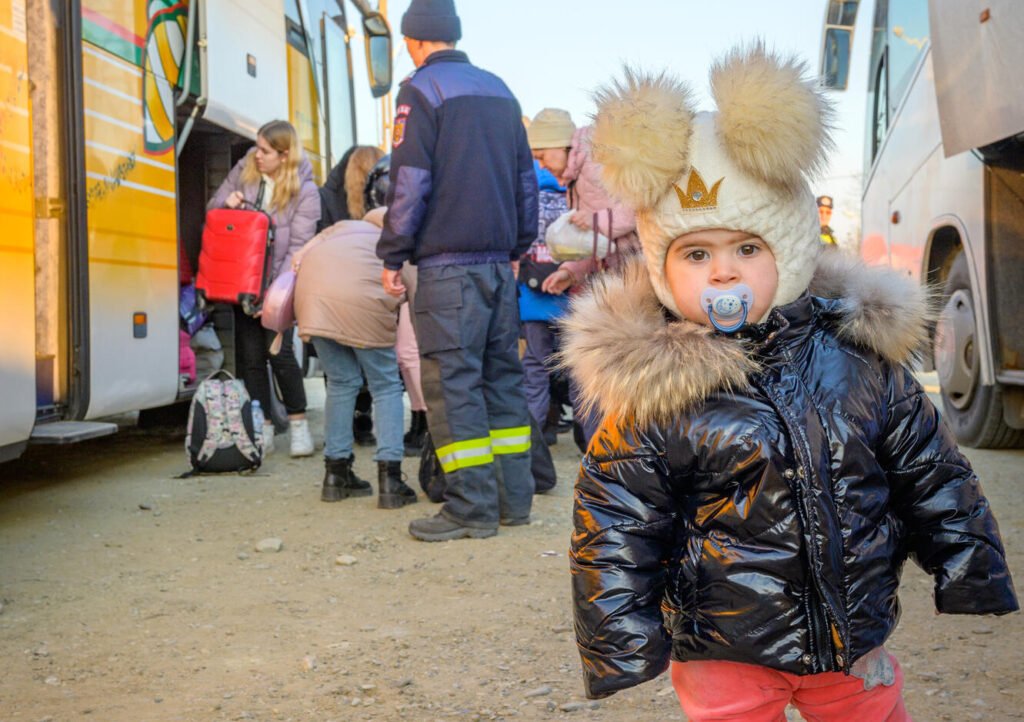 Ukrainalainen pikkutyttö, tutti suussa, seisoo kahden bussin välissä ja katsoo vakavana kameraan, tupsupipo päässä. Taustalla näkyy muita ihmisiä laittamassa tavaroita bussin tavaratilaan.