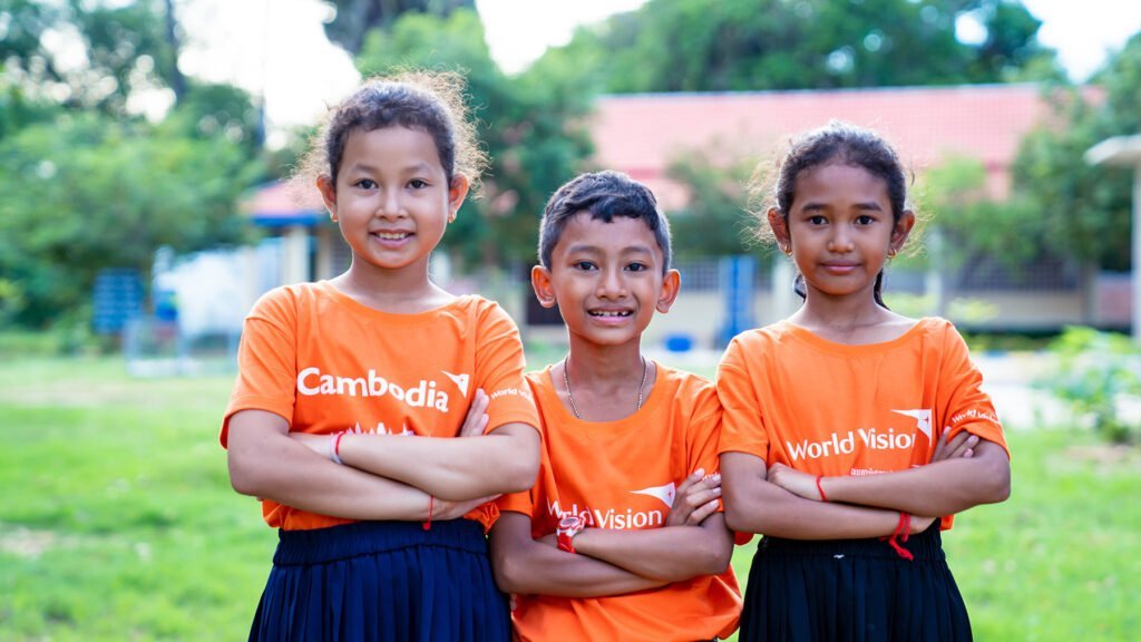 Kolme World Visionin oranssiin t-paitaan pukeutunutta kambodzalaista lasta poseeraavat hymyillen kameralle.