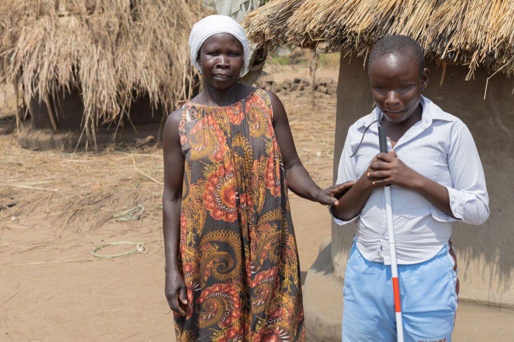 Rebecca Banye seisoo poikansa Hassanin kanssa kotinsa edustalla Adjumanin pakolaisalueella Ugandassa. Äiti koskettaa pojan käsivartta, pojalla on valkoinen sokeainkeppi kädessä.