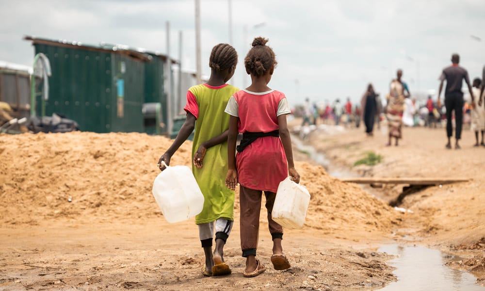 Kahta sudanilaista tyttöä kävelevät vierekkäin, vesikanisterit käsissä, keskellä pakolaisleiriä.