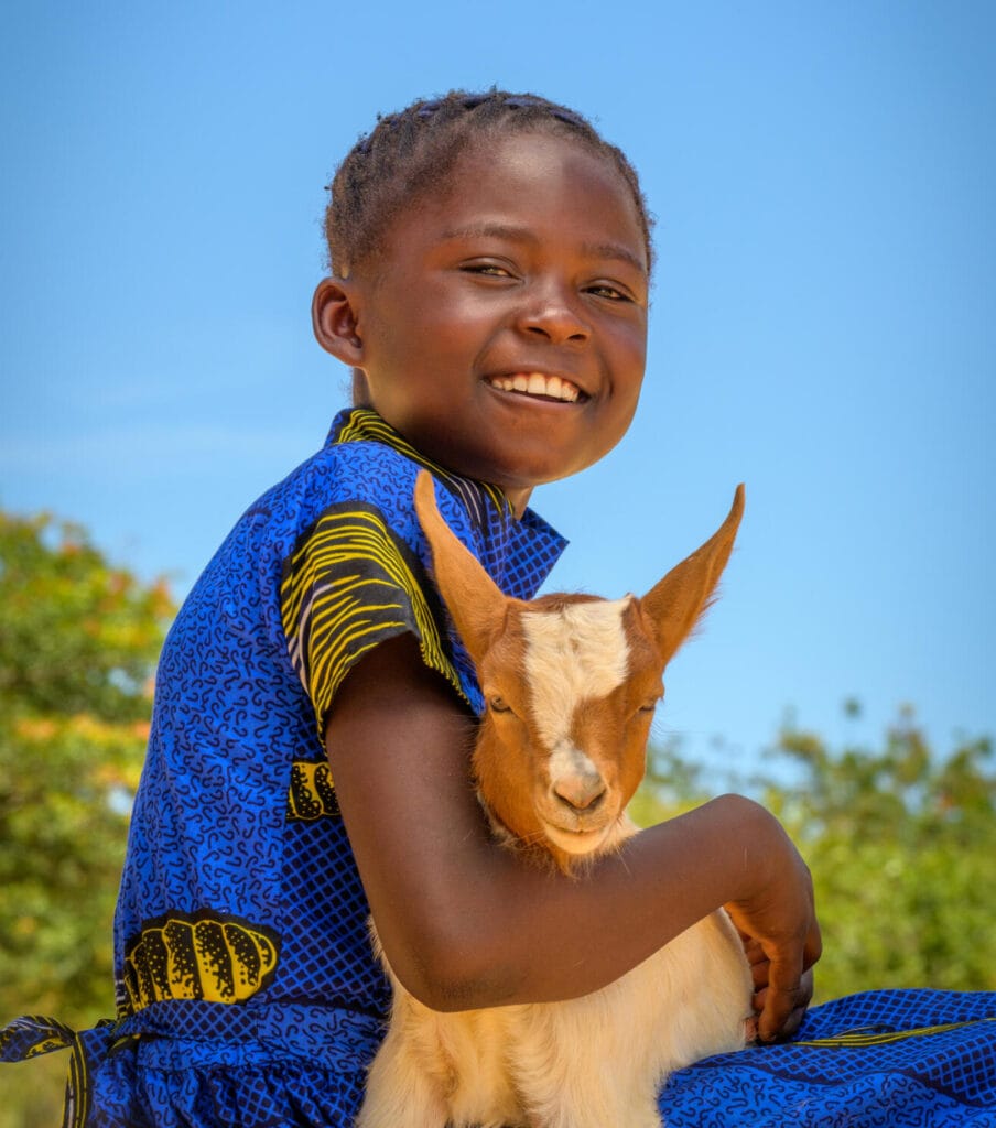 Tyttö hymyilee kameralle. Hän pitää sylissään vuohen kili.