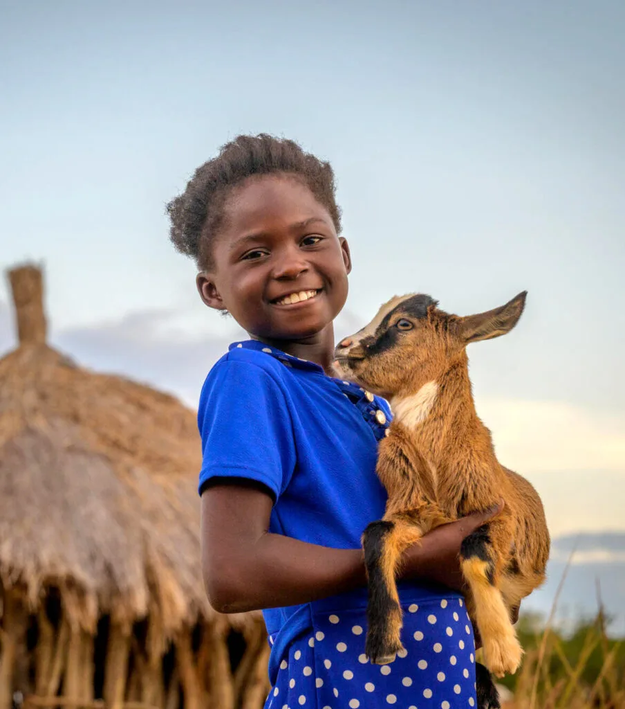 Tyttö hymyilee kameralle. Hän pitää sylissään vuohen kili.