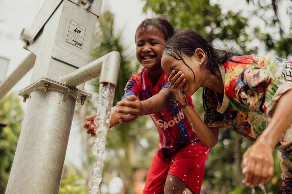 Kaksi kambodzalaista lasta pesevät kätensä ja kavonsa vesiputken kohdalla.