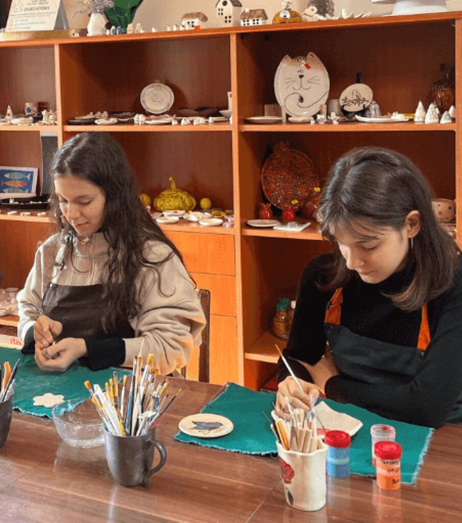 Sinipaitainen nainen seisoo ja muovailee keramiikkaa. Hänen edessään pöydän ääressä istuu naisen kaksi tytärtä maalaamassa ja työstämässä savea.