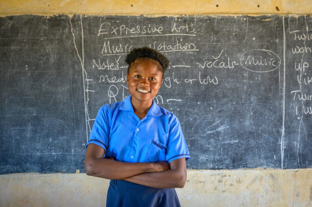 Sambialainen tyttö seisoo hymyilleen koulun taulun edessä.