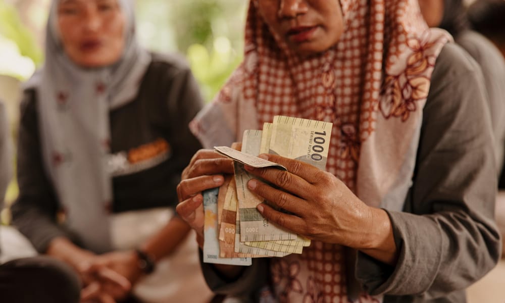 Lähikuva indonesialaisen naisen käsistä, jossa laskee seteleitä. Nainen on osa säästöryhmää ja hänen takana näkyy säästöryhmän muita naisia.