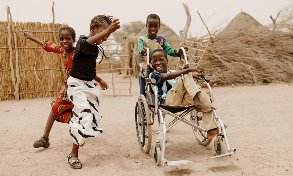 Senegalilainen poika istuu nauraen pyörätuolissa. Hänen ympärillä tanssi iloisesti kaksi nuorta tyttöä ja toinen poika pitää kiinni pyörätuolista.